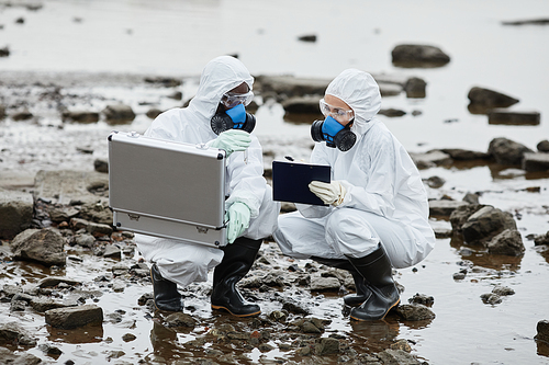 Two people wearing hazmat suits collecting samples by water, toxic waste and pollution concept