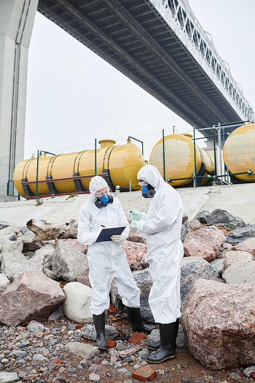 Full length portrait of two scientists wearing hazmat suits collecting samples outdoors, toxic waste and pollution concept