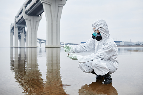 Side view full length portrait of scientist wearing hazmat suit collecting water samples, hands holding test tube, copy space