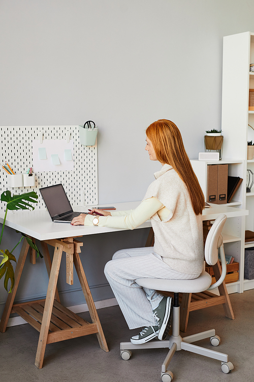 Vertical full length portrait of young red haired woman using laptop or studying at minimal home workplace