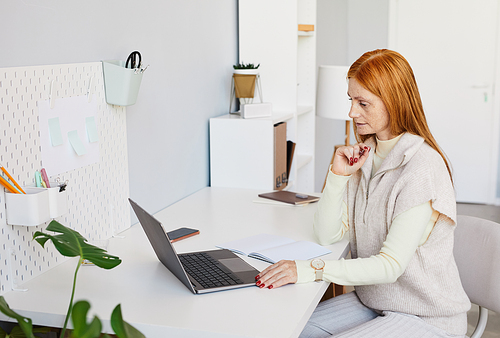 Side view portrait of red haired woman using laptop or studying at minimal home office workplace, copy space