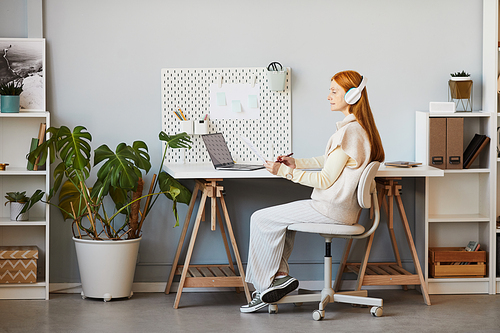 Full length portrait of red haired woman using laptop or studying at minimal home office workplace, copy space