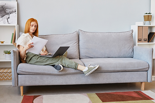 Full length portrait of adult red haired woman using laptop on sofa while working from home, copy space