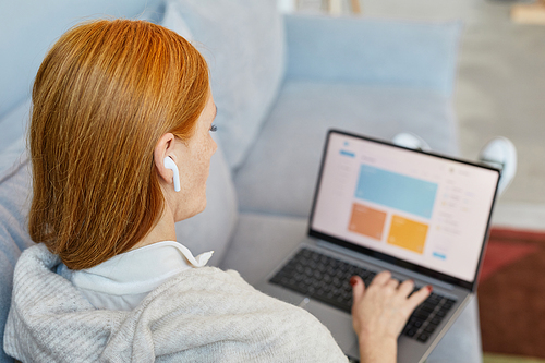 Back view portrait of adult red haired woman using laptop on sofa while studying online from home, copy space