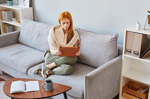 High angle portrait of adult red haired woman using digital tablet at home while sitting on sofa and studying online, copy space