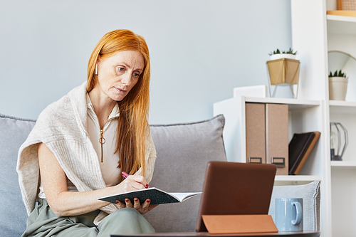 Portrait of adult red haired woman studying online at home and taking notes, copy space