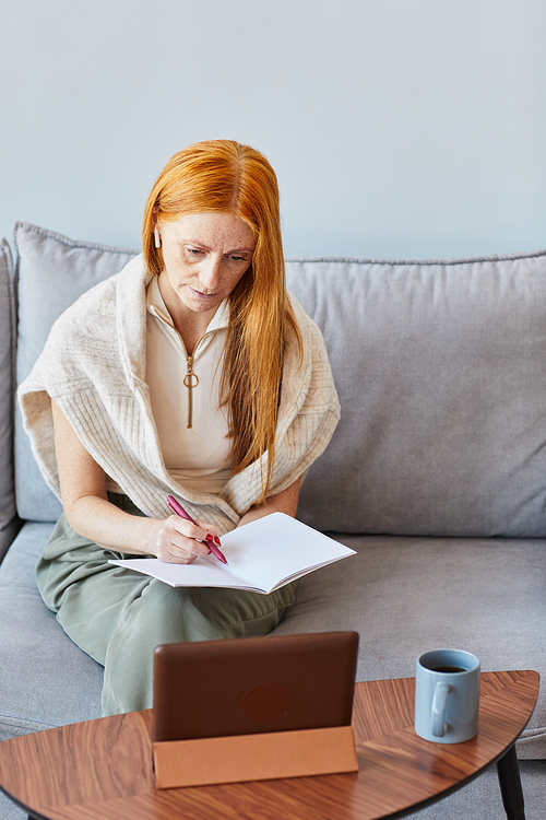 Vertical portrait of adult red haired woman studying online at home and taking notes