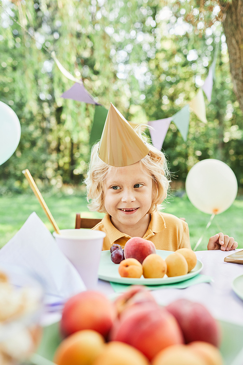 Vertical portrait of blonde boy wearing party hat at picnic table outdoors enjoying Birthday party in Summer