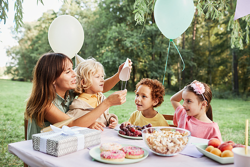 Portrait of young woman with son sitting at picnic table with group of kids during outdoor Birthday party in Summer