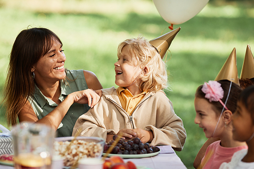 Portrait of happy young woman with son sitting at picnic table with group of kids during outdoor Birthday party in Summer