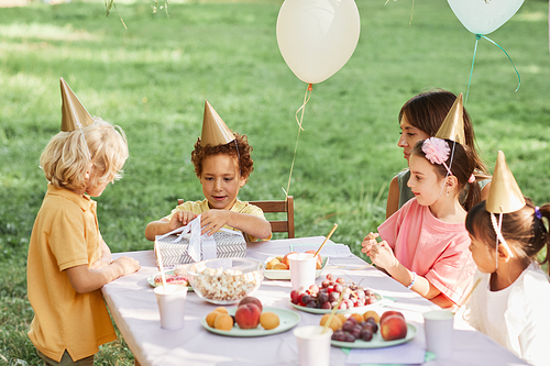 Portrait of cute curly boy opening gifts during outdoor Birthday party in Summer, copy space