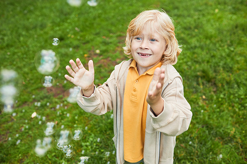 Portrait of cute blonde boy playing with bubbles outdoors in park and smiling, copy space