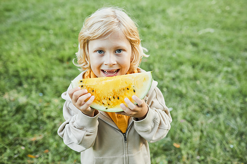 Waist up portrait of cute boy eating watermelon outdoors and smiling at camera, copy space