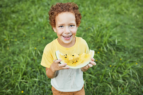High angle portrait of cute curly boy holding watermelon outdoors and smiling at camera, copy space