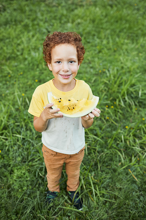 Vertical full length portrait of curly boy holding watermelon outdoors and smiling at camera