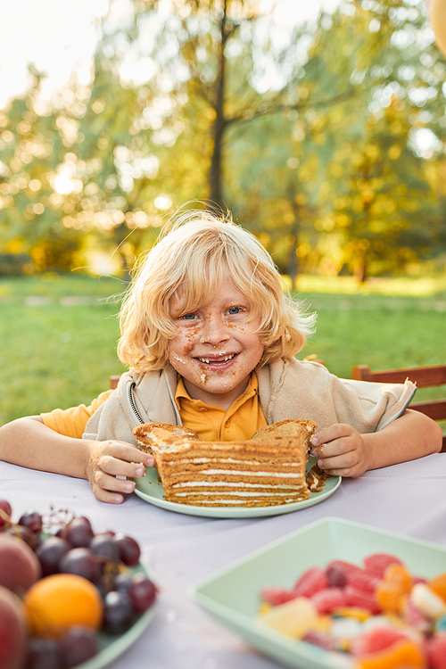 Vertical portrait of blonde boy eating cake during Birthday party outdoors and looking at camera