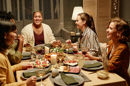 Group of happy friends sitting at dining table talking and laughing with each other at dinner party at home