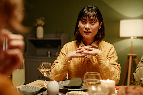 Asian young woman sitting at dining table and talking to her friends at dinner party