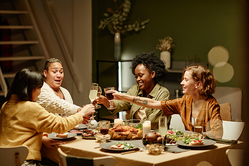 Group of young women toasting with glass of alcohol drinks and celebrating holiday together sitting at dining table at home