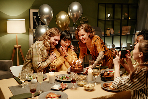 Happy young woman celebrating birthday with her friends at home, they sitting at dining table with birthday cake
