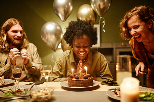 African happy woman sitting at the table with birthday cake with candles and laughing, she celebrating birthday with friends at home