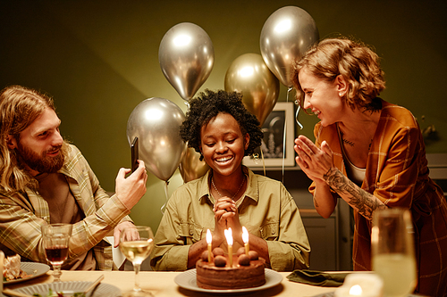 Happy African woman blowing candles on birthday cake while her friends congratulating her at dining table at home