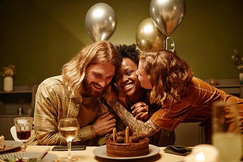 Young happy couple congratulating their friend with birthday, they sitting at the table kissing her and embracing