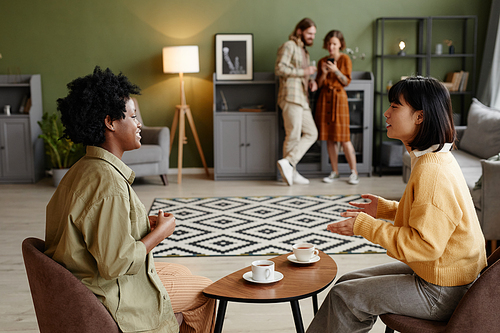 Young women sitting at coffee table drinking coffee and talking to each other during home party with couple in the background