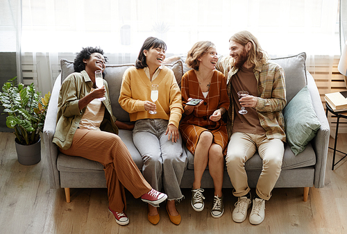 Group of happy friends sitting on sofa drinking champagne and laughing during conversation in the room