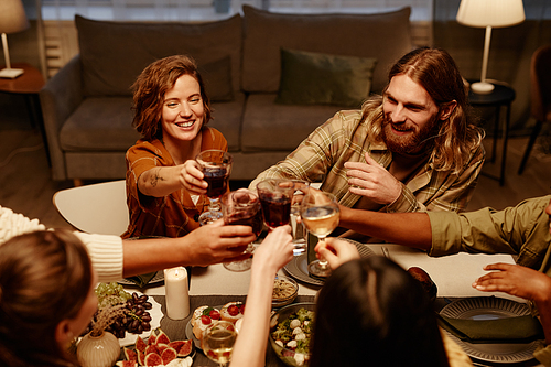 Group of happy friends toasting with glasses of red wine and congratulating each other sitting at dining table at home