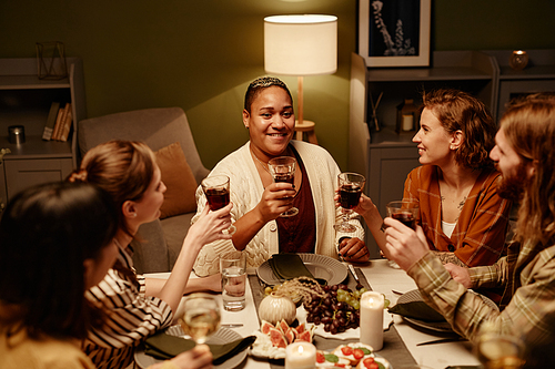 Group of young people holding glasses with red wine and celebrating the holiday while sitting at dining table at home
