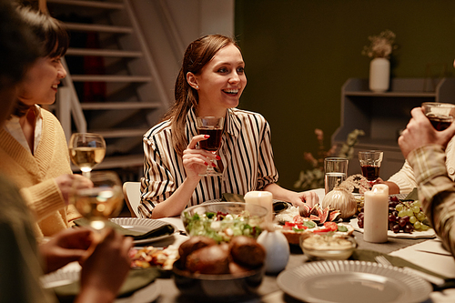 Happy young woman drinking wine and talking to her friends at dining table during dinner party at home