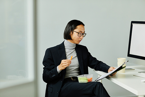 Minimal portrait of Asian businessman eating healthy snacks while reading documents in office