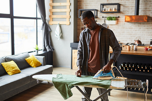 Portrait of young African-American man ironing clothes in studio apartment, bachelor lifestyle and household chores concept, copy space