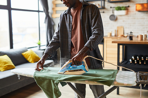 Cropped shot of young African-American man ironing clothes in studio apartment, bachelor lifestyle and household chores concept, copy space
