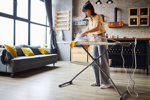 Full length portrait of young Asian woman ironing clothes at home with steam, household chores concept, copy space