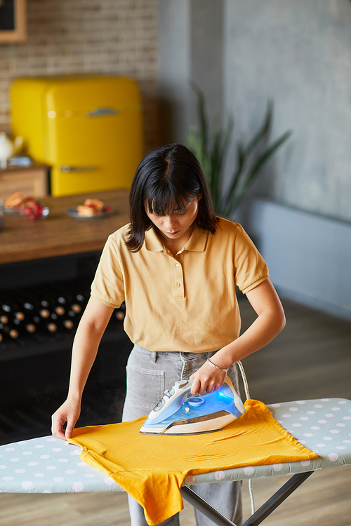 Vertical portrait of young Asian woman ironing clothes at home and doing household chores
