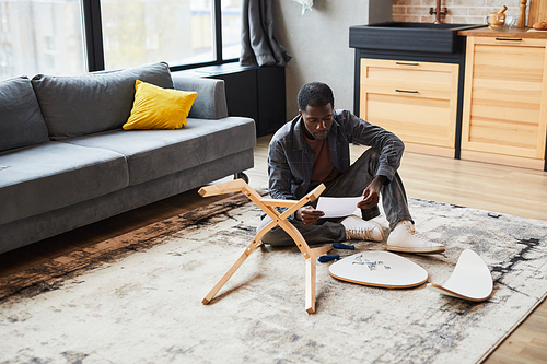 Wide angle portrait of young African-American man assembling furniture at home, copy space