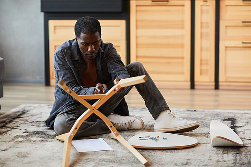 Full length portrait of young African-American man assembling furniture at home, copy space