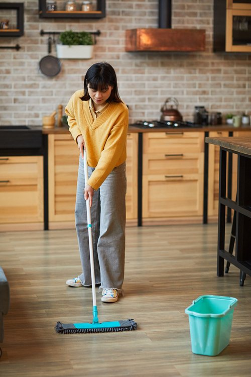 Vertical full length portrait of young Asian woman mopping floors while cleaning cozy apartment