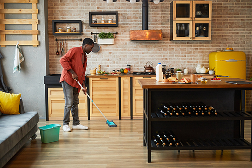 Wide angle view at young African-American man mopping floors while cleaning cozy apartment, copy space