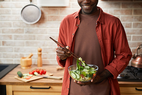 Cropped shot of smiling black man mixing salad in glass bowl while enjoying healthy meal in cozy kitchen, copy space