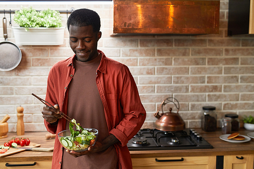 Waist up portrait of smiling black man mixing salad in glass bowl while enjoying healthy meal in cozy kitchen, copy space