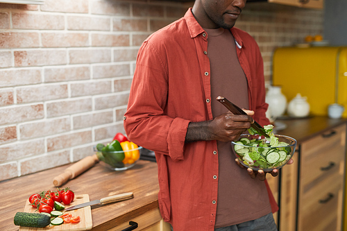 Cropped shot of young black man mixing salad in glass bowl while enjoying healthy meal in cozy kitchen
