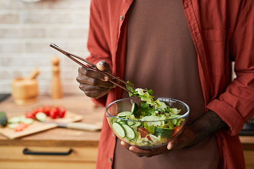 Close up of young black man mixing salad in glass bowl while enjoying healthy meal in cozy kitchen, copy space