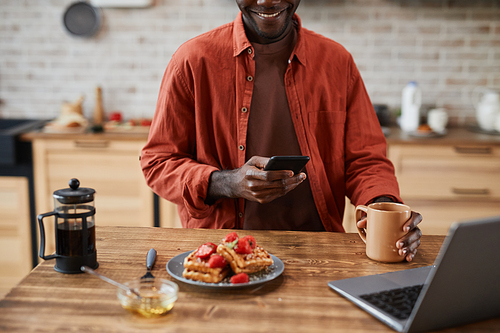 Cropped portrait of black young man using smartphone while enjoying breakfast at home kitchen, copy space