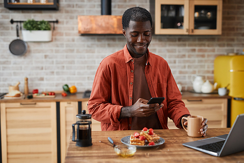 Portrait of smiling black man using smartphone while enjoying breakfast at cozy home kitchen, copy space