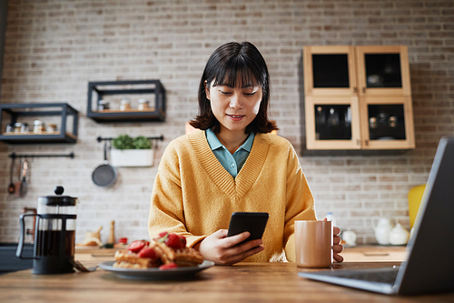 Portrait of smiling Asian woman using smartphone while enjoying breakfast at cozy home kitchen, copy space