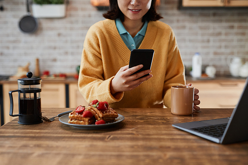 Cropped shot of smiling Asian woman using smartphone while enjoying breakfast at cozy home kitchen, copy space