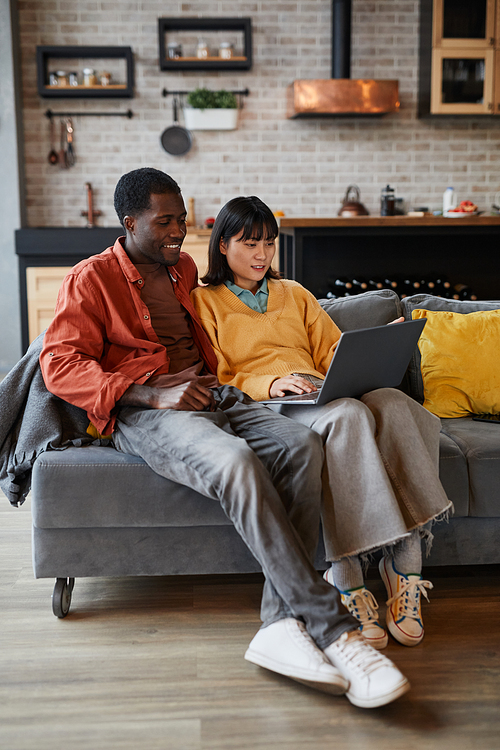 Vertical full length portrait of young couple using laptop while relaxing on couch in cozy home interior